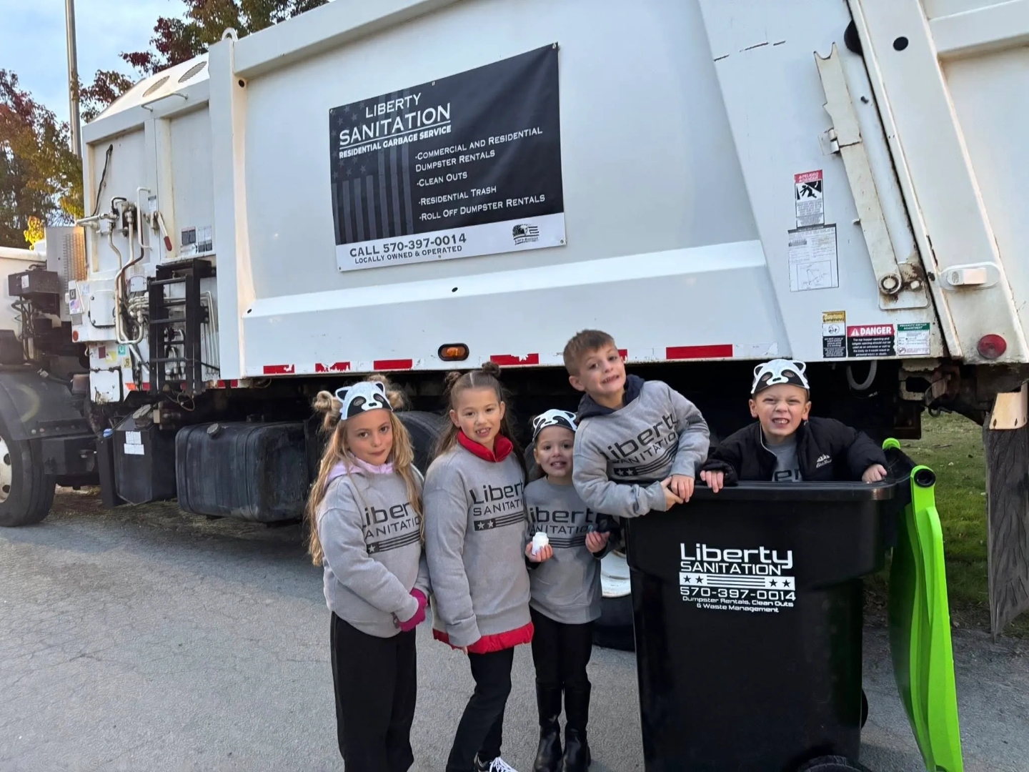 Children wearing Liberty Sanitation hoodies standing beside a branded cart and Liberty garbage truck